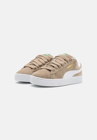 Baskets en daim marron clair avec semelle en caoutchouc blanche. Comprennent des lacets ronds, une étiquette verte sur la languette et une bande blanche sur le côté. Détail du logo doré.