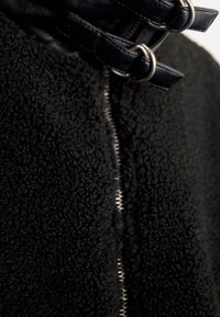 Schwarze Fleecejacke mit einer strukturierten, weichen Außenseite, einem zentralen Reißverschluss und schwarzen Lederelementen mit Metallbeschlägen.
