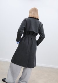 Charcoal gray wool coat with a belted waist, collar, and layered back design. The model wears gray trousers and black shoes.