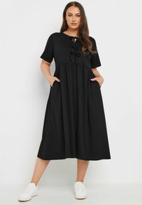 Musta midi-mekko pehmeästä kankaasta, jossa on lyhyet hihat, vyötärö on kiristetty, siinä on kaksi sivutaskua ja etuosassa solmimisdetalji. Yhdistetty valkoisiin lenkkareihin.