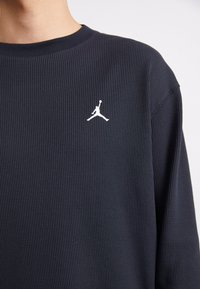 Maglietta a maniche lunghe blu navy con una trama a punto waffle. Presenta un logo Jumpman bianco sul lato sinistro del petto. Collo tondo.