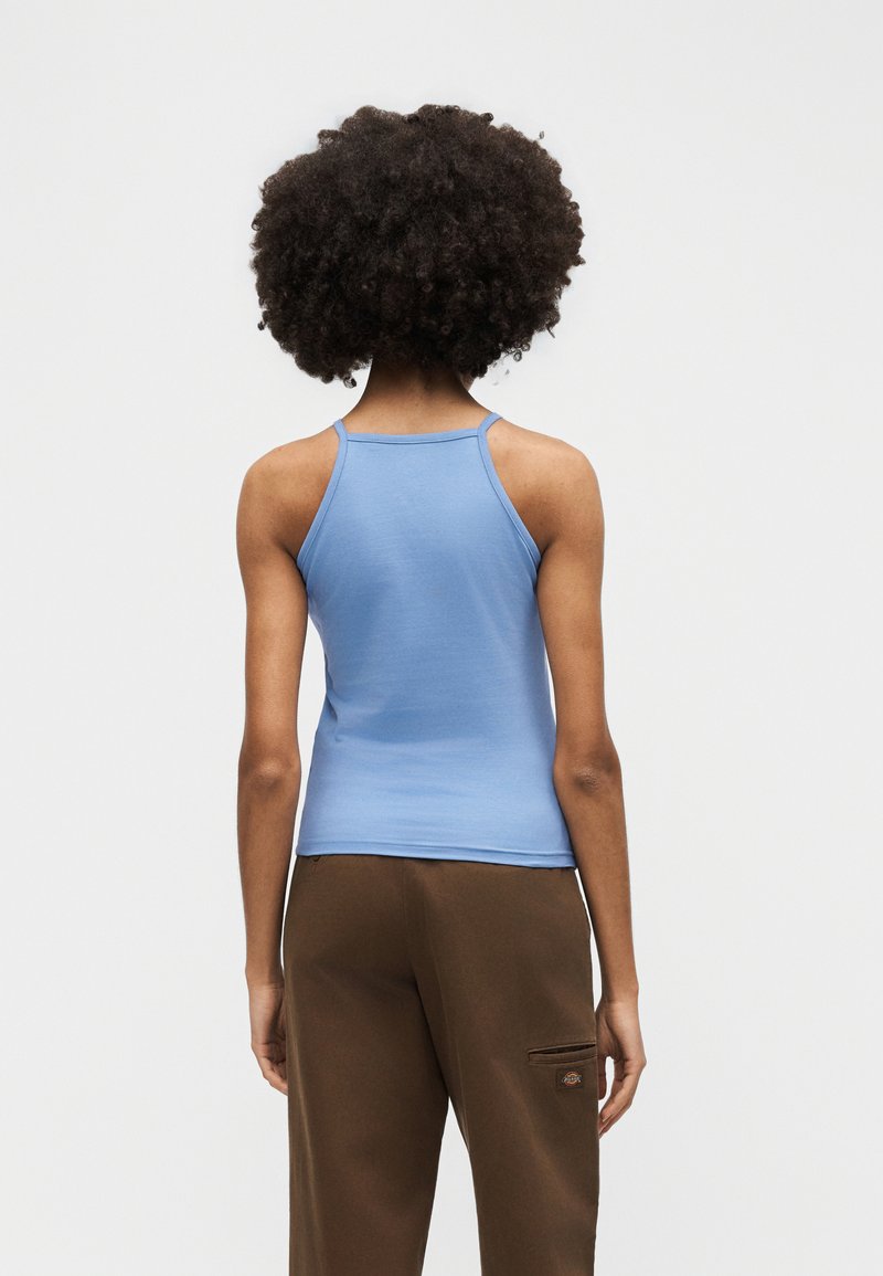 Persona con capelli ricci che indossa una canottiera azzurro chiaro e pantaloni marroni, vista di spalle su uno sfondo bianco uniforme.