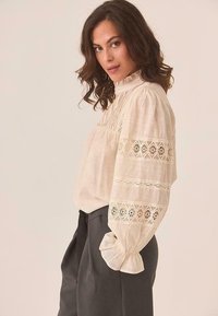 Blusa de color beige claro con mangas largas abullonadas, tela texturizada, ribete de encaje con patrones geométricos, cuello alto y corte holgado.