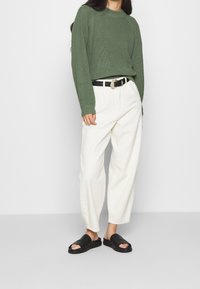 Pull côtelé vert, pantalon blanc à jambes larges, ceinture noire à boucle argentée et sandales noires à enfiler. Tenue simple et décontractée.