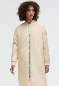 Heller beiger langer Mantel mit einem Reißverschluss, seitlichen Taschen, gerippten Bündchen und einer glatten, glänzenden Textur. Ideal für kühles Wetter.