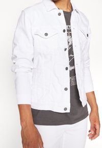 Chaqueta de denim blanca con cierre de botones en la parte delantera, dos bolsillos en el pecho y un diseño ligeramente ajustado. Presenta botones oscuros y mangas largas.