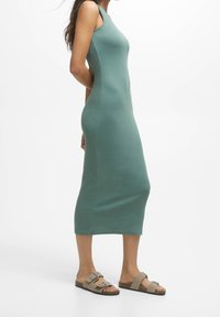 Vestido midi sin mangas de tejido verde claro, diseño ajustado, textura suave, combinado con sandalias planas beige que presentan dos hebillas.