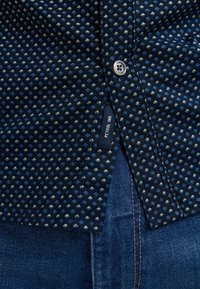 Maglietta navy con un motivo a rombi in azzurro chiaro e crema. Dettaglio dei bottoni visibile. L'etichetta riporta "Petrol Ind." Jeans parzialmente visibili sotto.