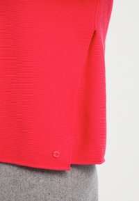 Czerwony sweter z prążkowanej dzianiny z bocznymi rozporkami, z małym detalem w postaci guzika z logo na dolnej krawędzi. Miękka faktura i intensywny kolor.