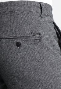 Grijze broeken met een visgraatpatroon, voorzien van een zwarte knoop, met een achterzak en een klein merklabel op de tailleband.