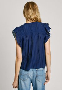 Top senza maniche blu navy con dettagli arricciati sulle spalle, tessuto testurizzato e design sul retro arricciato, abbinato a jeans azzurri.