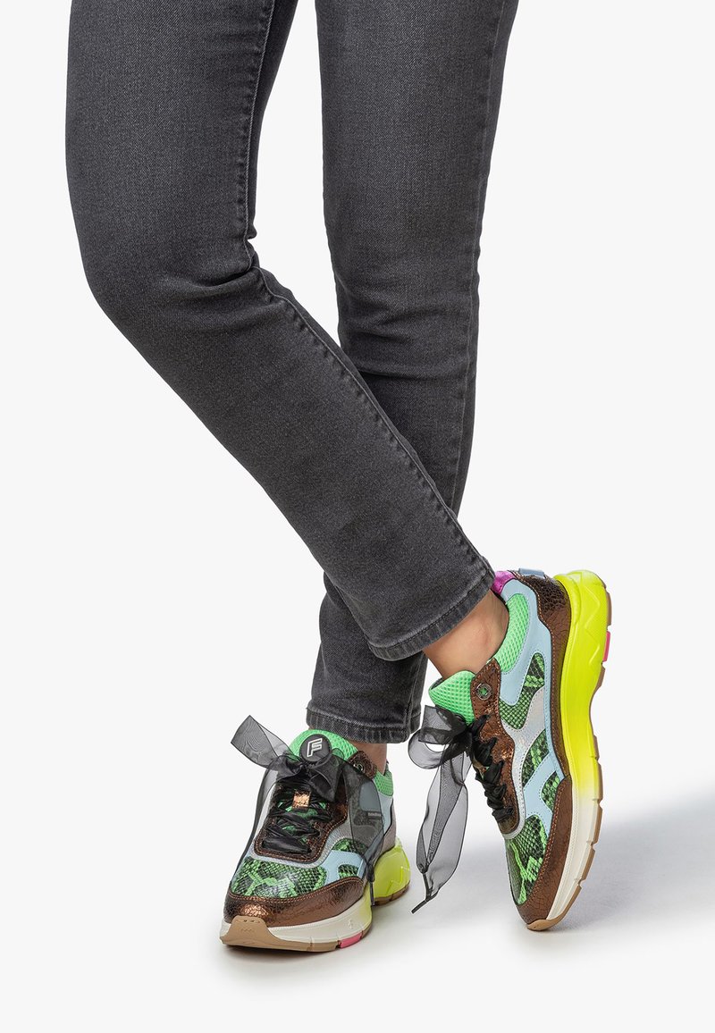 Nogawki w ciemnoszarych dżinsach skrzyżowane, w sportowych butach z neonowozieloną siateczką, brązowymi i pastelowoniebieskimi detalami, czarnymi przezroczystymi sznurowadłami oraz jasnolicymi podeszwami.