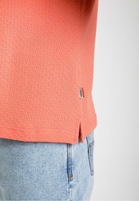 Korallitooni tekstuuriga särk, millel on mustriline viimistlus, külgmised lõhikud ja väike silt. Kombineeritud heledate siniste denim-lühikeste pükstega.