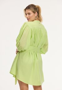 Donna con capelli biondi raccolti in una coda di cavallo, che indossa grandi orecchini d'oro e un vestito largo, verde chiaro, con cintura, che guarda oltre la spalla.