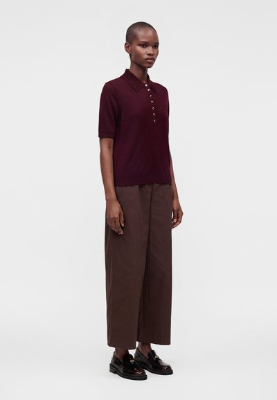 Femme debout portant un polo manches courtes bordeaux foncé, un pantalon large marron et des mocassins noirs sur un fond blanc uni.