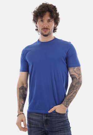 Uomo con capelli ricci e tatuaggi su entrambe le braccia, indossa una maglietta blu e jeans, in piedi con una mano in tasca.