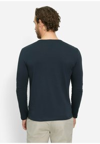 Mann mit lockigem Haar, der ein dunkles Langarmshirt und beige Hosen trägt, steht mit dem Rücken zu einem schlichten weißen Hintergrund.