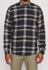 Koszula w kratę w kolorze granatowym, białym i czerwonym. Posiada kołnierzyk zapinany na guziki, długie rękawy i kieszeń z przodu. Wykonana z teksturowanej mieszanki bawełny.