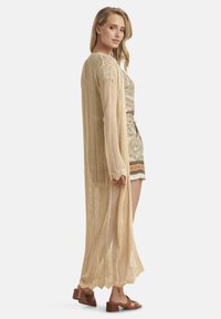 Beige gestrickte Strickjacke mit offenem Muster, langen Ärmeln und seitlichen Schlitzen, kombiniert mit einem gemusterten Kleid und braunen Blockabsatzsandalen.