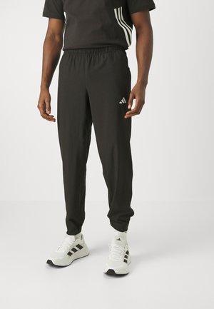 Mann trägt schwarze Adidas-Trainingshose, schwarzes Shirt mit weißen Streifen, weiße Adidas-Socken und weiße Sneakers, steht auf einfarbigem Hintergrund.