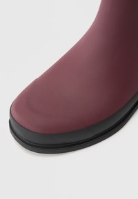 Botte de pluie en caoutchouc bourgogne avec une finition mate lisse, un bout rond et une semelle noire. Le design présente une tige sans coutures et une construction robuste.