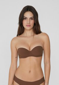 Brązowy stanik bez ramiączek z formowanymi miseczkami i gładkiego materiału; bezszwowy design zapewnia smukłą sylwetkę. Modelka ma długie brązowe włosy i tatuaż.