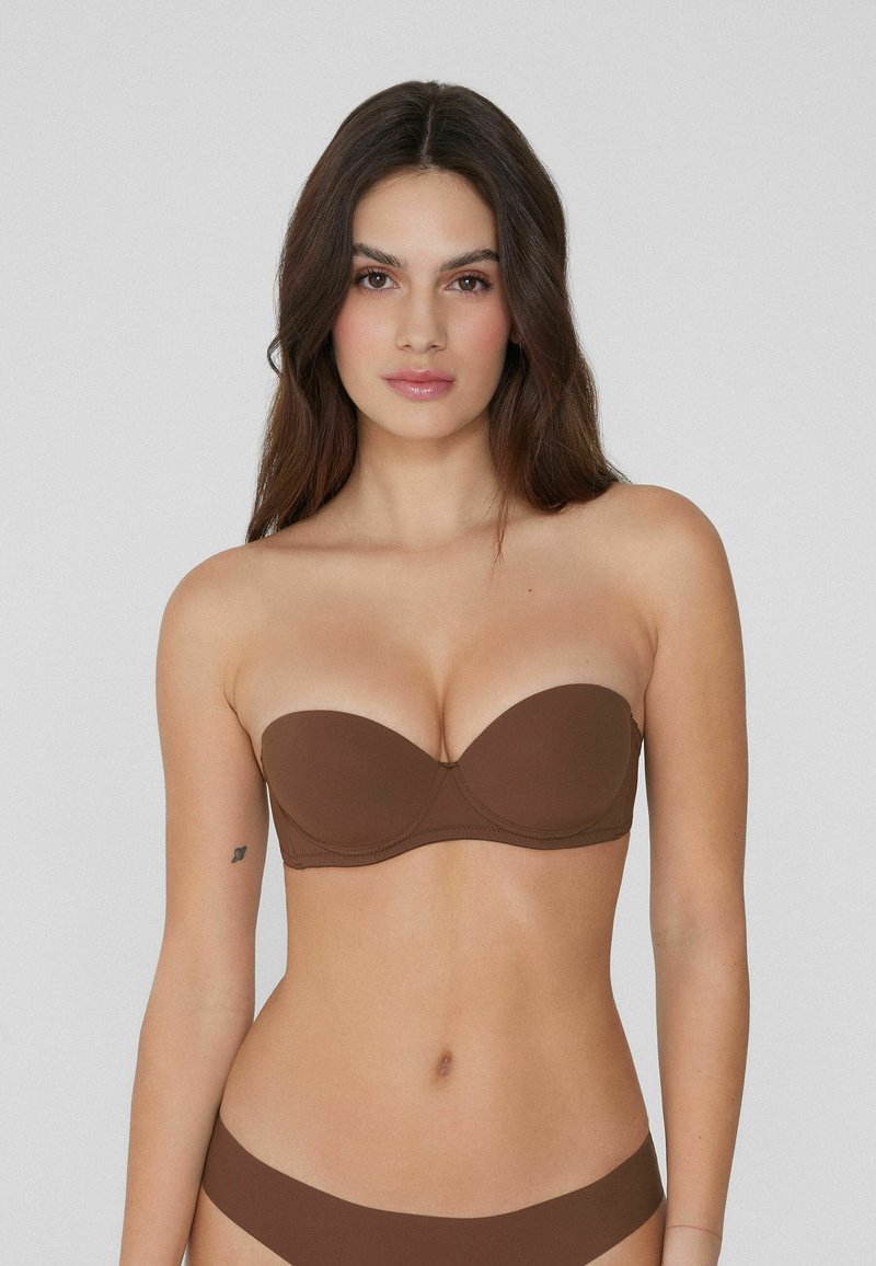 Brązowy stanik bez ramiączek z formowanymi miseczkami i gładkiego materiału; bezszwowy design zapewnia smukłą sylwetkę. Modelka ma długie brązowe włosy i tatuaż.