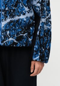 Μανίκι μπουφάν από φλις με σχέδιο σε μπλε, μαύρο και ανοιχτό γκρι, με υφή υφάσματος και ορατό πλαϊνό ραφτό, επάνω σε μαύρο παντελόνι.