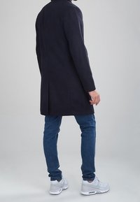 Cappotto in misto lana blu navy con taglio dritto, spacco posteriore e collo alto, abbinato a jeans blu e sneakers bianche, mostrato da dietro.