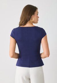 Femme aux cheveux bruns portant des boucles d'oreilles en or, un haut ajusté à manches courtes bleu marine et un pantalon blanc, de dos contre un fond uni.