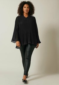 Schwarze plissierte Bluse mit V-Ausschnitt und weiten Glockenärmeln, kombiniert mit eleganten schwarzen Leggings und glänzenden schwarzen Schuhen.