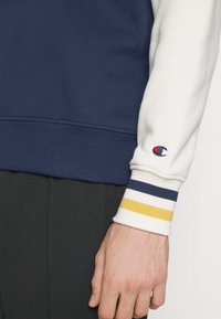 Tmavě modrá a krémová mikina s proužkovanými žebrovanými manžetami; na rukávu je vyšívané logo; měkká látka s volným střihem.