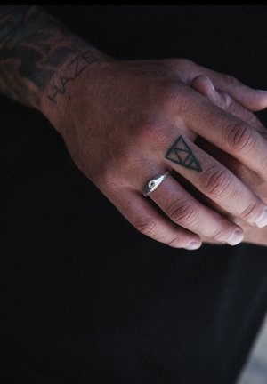Silver ring with a prominent eye design rests on a hand featuring multiple tattoos. The hand has a textured appearance and neutral skin tone.