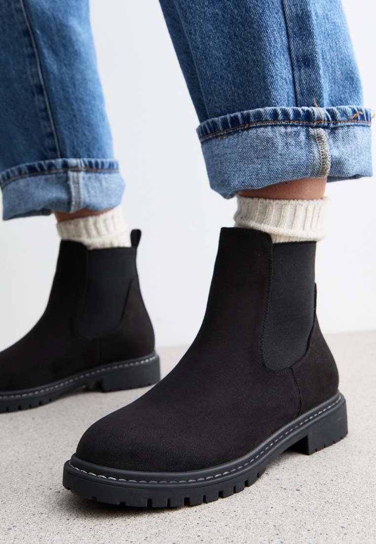 Schwarze Wildleder-Stiefeletten mit kräftigen Sohlen, kombiniert mit cremefarbenen Socken und hochgekrempelten blauen Jeans auf einem hellgrauen Boden.