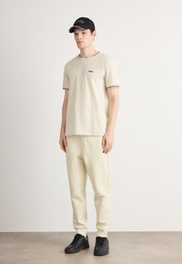 T-shirt beige à manches courtes avec des bordures noires et un petit logo, assorti à un pantalon de survêtement beige. Baskets noires avec une surface texturée.