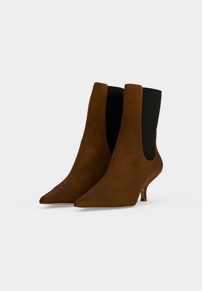 Paire de bottines en daim marron à bouts pointus, panneaux latéraux élastiques noirs et talons courts et fins sur fond clair.