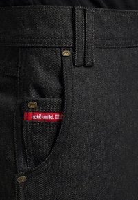 Zwarte denim jeans met een gestructureerde afwerking, voorzien van een gouden metalen knop en een rood-wit label op de voorzak.