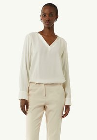 Weiße Bluse mit langen Ärmeln und V-Ausschnitt, aus glattem Stoff. Kombiniert mit beigen, maßgeschneiderten Hosen. Einfaches Design, neutrale Farbpalette.