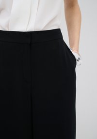 Zwarte broeken van gladde stof, met zijzakken en een strakke tailleband. In combinatie met een witte blouse die aan de bovenkant zichtbaar is.
