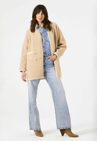 Beige fleece jas met een ontspannen silhouet, voorzien van dubbele knopen, crème accenten en zijzakken, gelaagd over een denim jumpsuit.