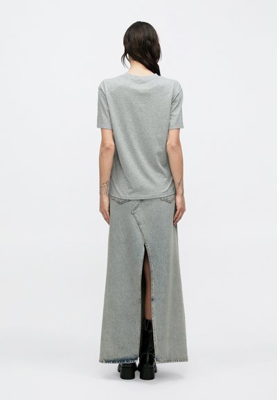 Pikkade tumedate juustega naine, kes kannab halli lühikeste varrukatega t-särki, helefarkudest maksiseelikut tagumise lõhikuga ja mustasid pahkluu saapaid, vaate suunaga selja poole.