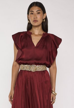 Vrouw met lang donker haar die een bordeaux satijnen jurk met geruite mouwen draagt, een geweven beige riem, gouden oorringen, armbanden en ringen.