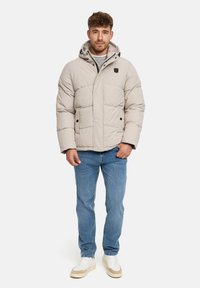 Beige dunjakke med lynlås, store lommer og justerbar hætte. Kombination med lyseblå jeans og hvide sneakers med beige detaljer.