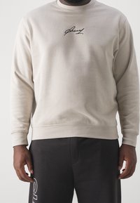 Felpa beige in tessuto morbido con maniche lunghe e scollo rotondo. Ricamo nero con firma distintiva sul petto.