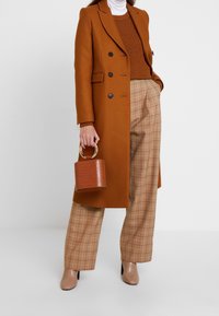 Brown double-breasted coat, knit sweater, beige plaid pants, nude heeled shoes, and small textured handbag with a circular handle.