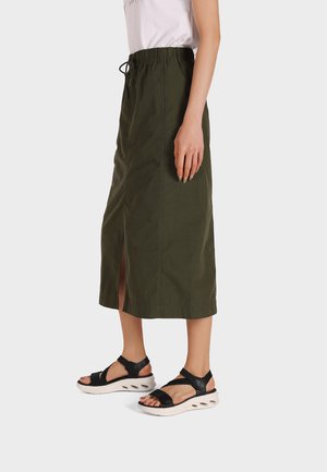 Donna che indossa una gonna midi verde oliva con spacco laterale, top bianco e sandali neri con cinturino e suole bianche spesse, in piedi di profilo.