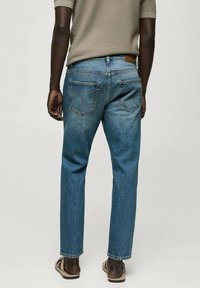 Mann in hellblauen geraden Jeans, beigem kurzärmligen Hemd und beigen Sandalen, mit dem Rücken zur Kamera stehend.