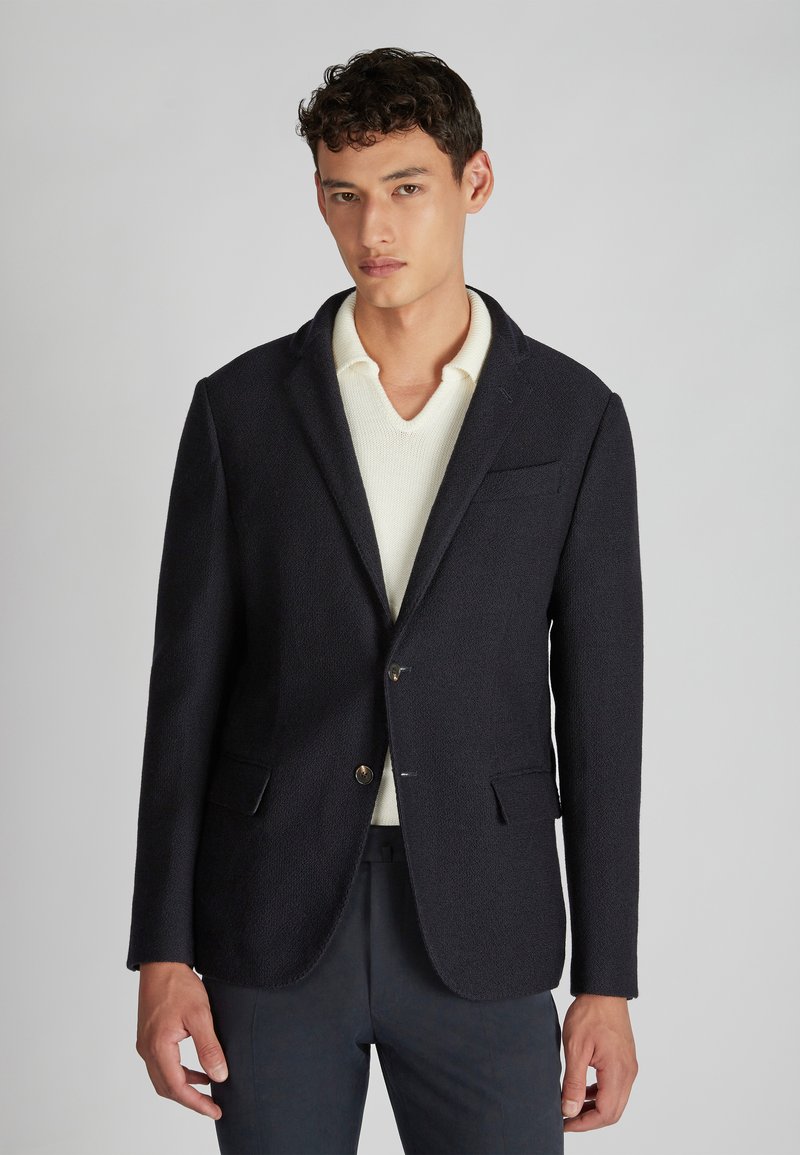 Granatowa marynarka o fakturze z zapięciem na jeden guzik, z klapami w kształcie notcha i dwiema przednimi kieszeniami, noszona na kremowym swetrze z dekoltem w serek.