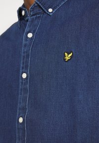 Camicia in denim blu scuro con colletto a punta. Presenta bottoni bianchi e un piccolo logo di un uccello ricamato in giallo vicino al petto.