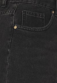 Vaqueros de mezclilla negra con una textura suave, que presentan un pequeño bolsillo frontal y acentos metálicos en tono dorado. Se puede ver un distintivo grosor en las costuras.